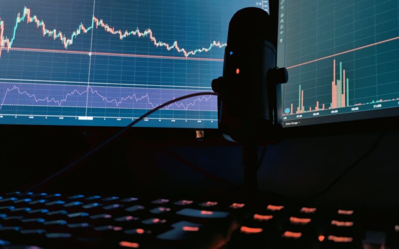A display of two cryptocurrency markets on different monitors with a microphone and multicoloured keyboard in the foreground.